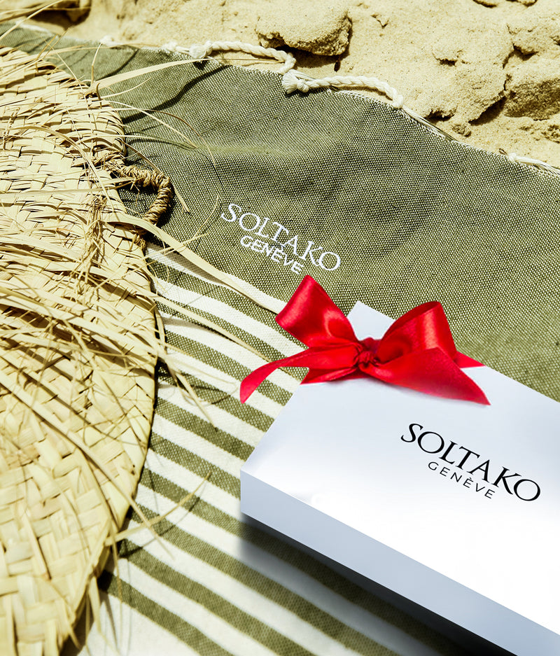 Strandtuch in Khaki grün Farben mit weißen Streifen im Sand liegend und SOLTAKO Geschenkbox mit roter Schleife