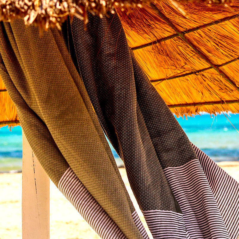 ein khakifarbenes und ein graues Strandtuch die an einem Sonnenschirm am Strand hängen