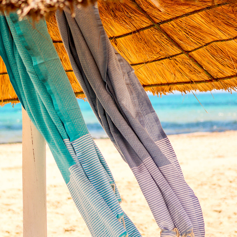 ein aquablaues Fouta und ein graues Strandtuch unter einem Sonnenschirm am Strand