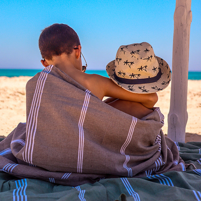 zwei kleine Brüder in beiger Fouta die auf einem khakifarbenen Strandtuch am Strand sitzen 