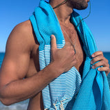 ein Mann am Strand der eine blaue Saunatuch Fouta auf den Schultern trägt