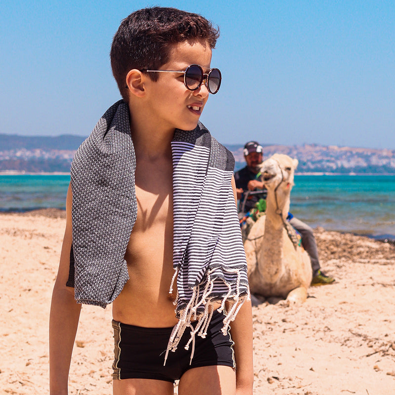 ein Kind mit Sonnenbrille und dunkelgrauem Strandtuch auf den Schultern am Strand
