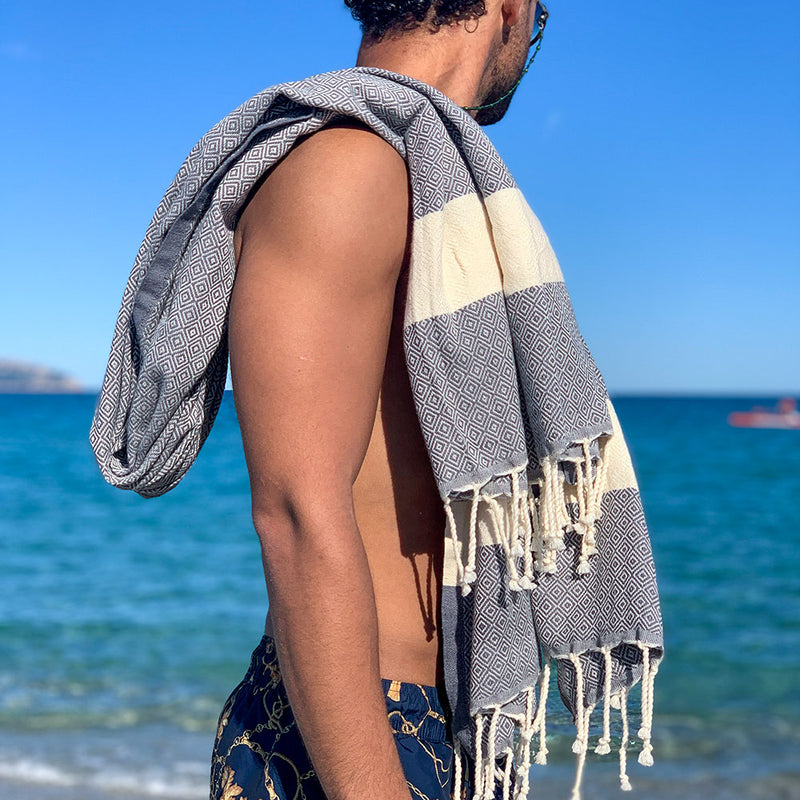 Mann in kurzen Hosen mit grauem Strandtuch auf den Schultern am Strand