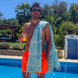 ein Mann am Pool mit einem Glas Saft in der Hand und einem aquablauen Strandtuch auf den Schultern