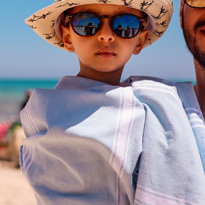 ein Kind mit Sonnenbrille und Hut das ein hellblaues Strandtuch trägt 