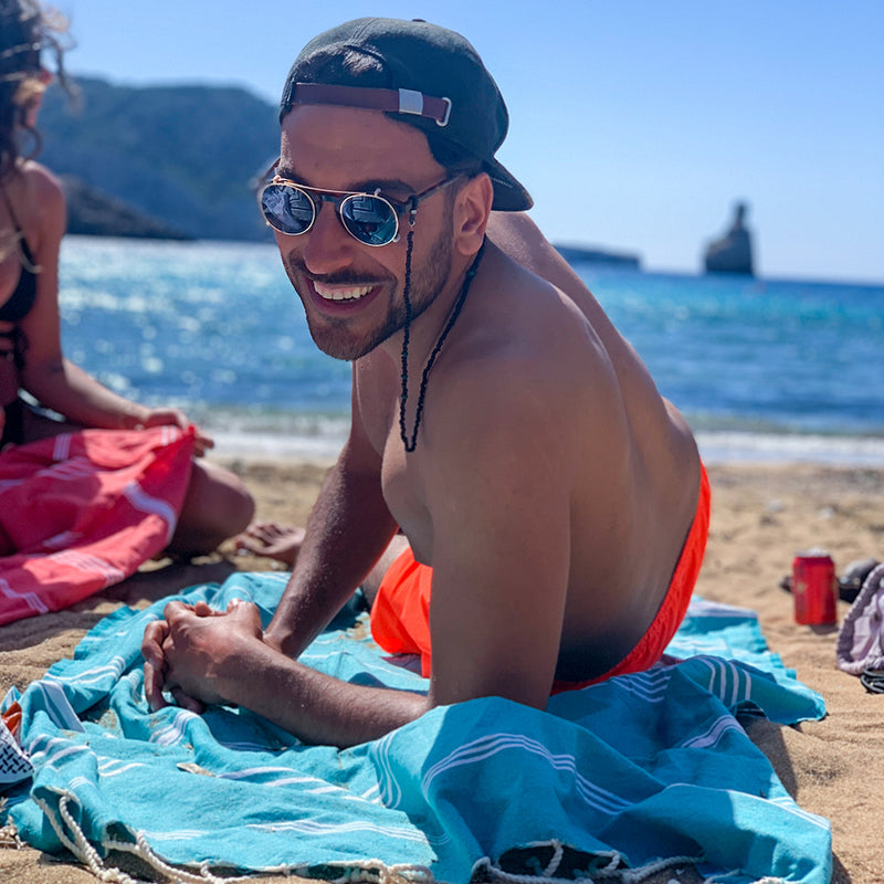 Mann mit Sonnenbrille auf einer aquablauen Fouta am Strand liegend