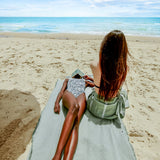 zwei Mädchen am Strand eine liegt auf einem khakifarbenen Strandtuch die andere trägt ein khakifarbenes fouta